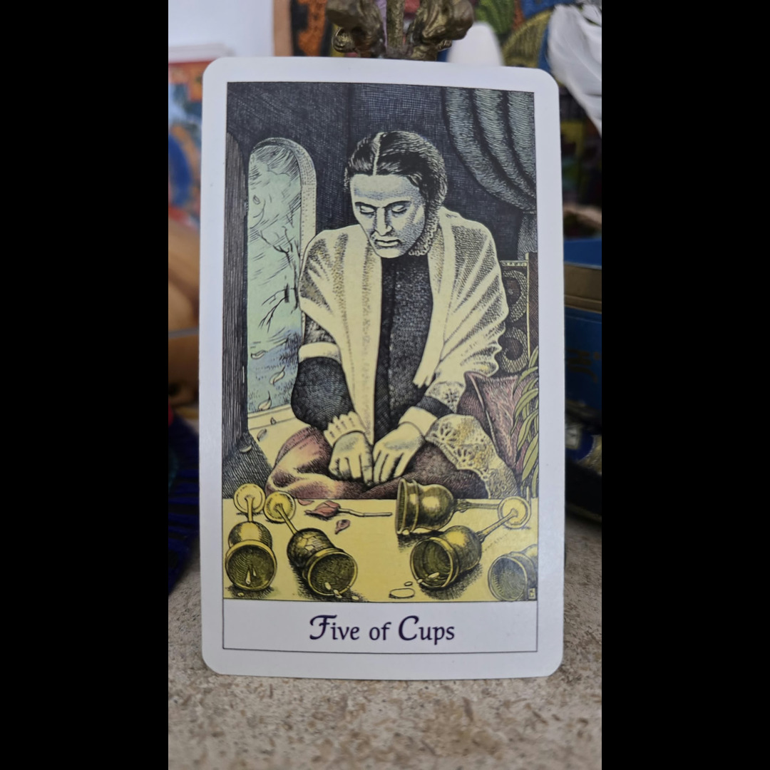 White-bordered Five of Cups tarot card showing a seated woman in a cream shawl beside a mirror, with five golden cups tipped on a yellow ground.