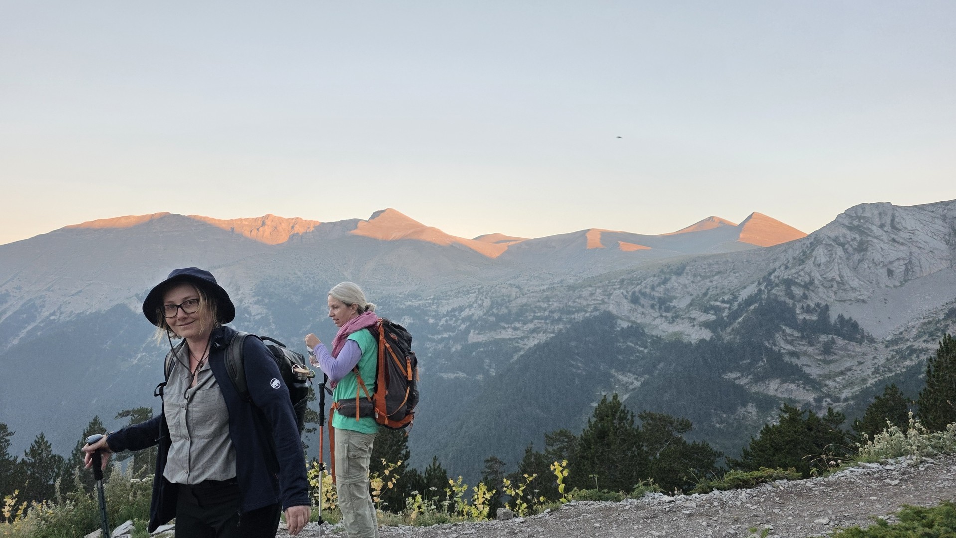 Two hikers on Mount Olympus with backpacks and walking poles, with evening light on the mountain peaks behind them.