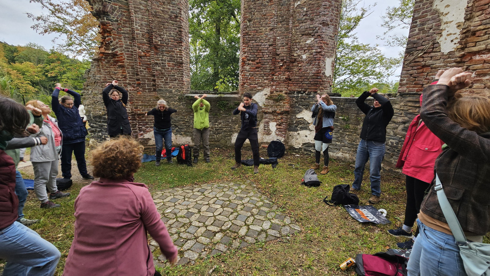 People in casual clothes stand in a circle with arms raised in front of tall brick abbey ruins, bags on the grass.