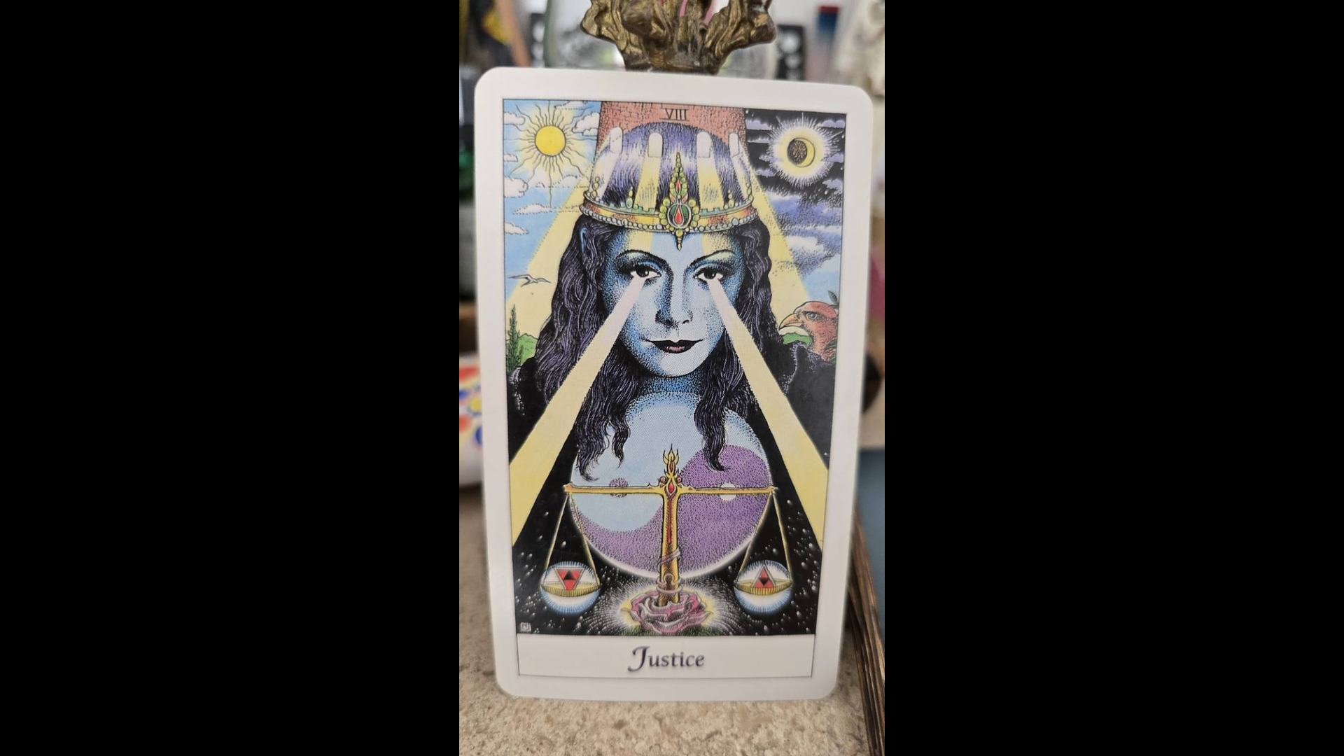 A tarot card titled Justice, numbered VIII, showing a blue-skinned crowned figure holding scales, propped upright on a speckled table