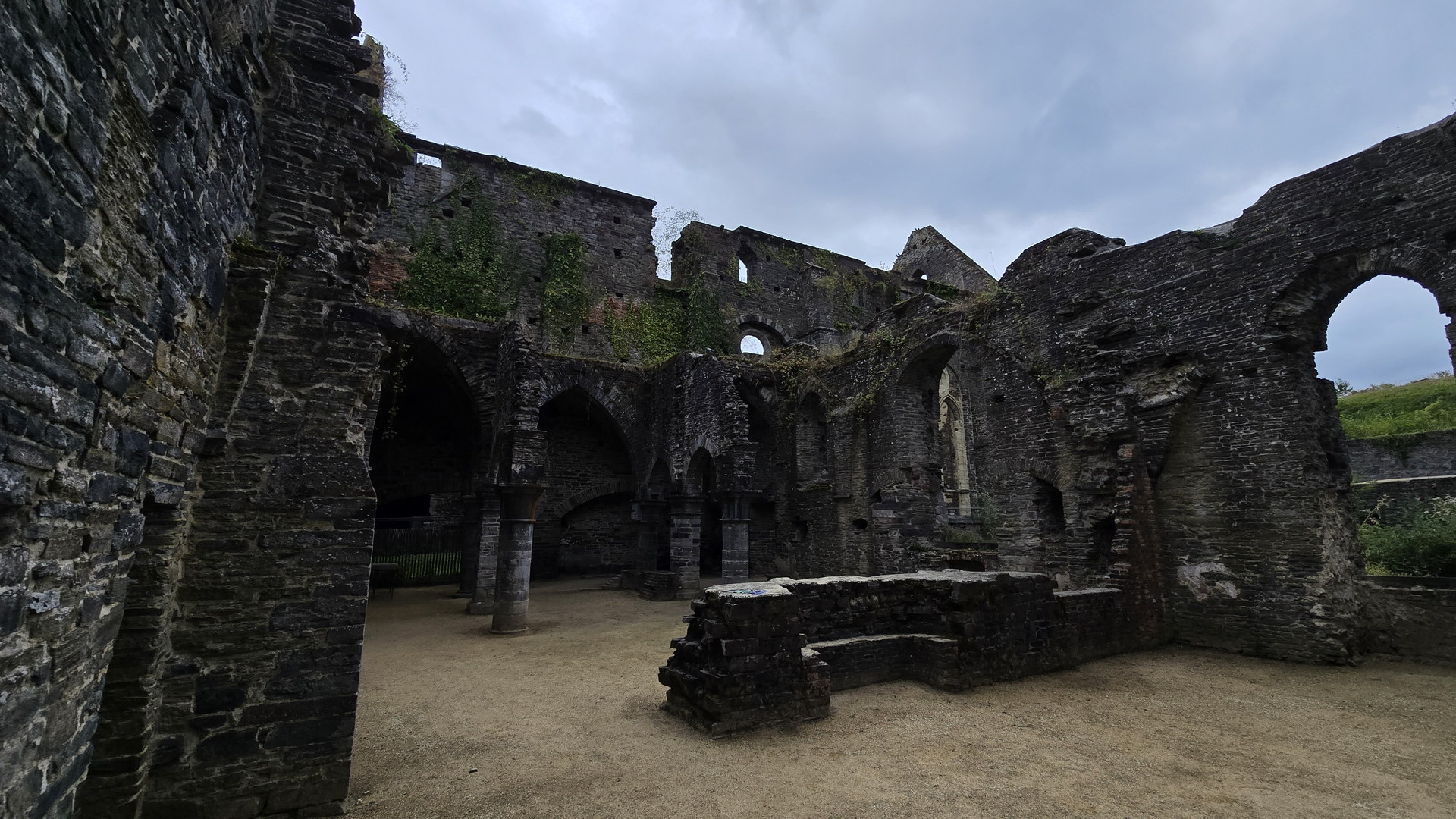Interior of the ruined Villers-la-Ville abbey: tall grey stone walls, gothic arches, ivy, sandy floor and overcast sky above.