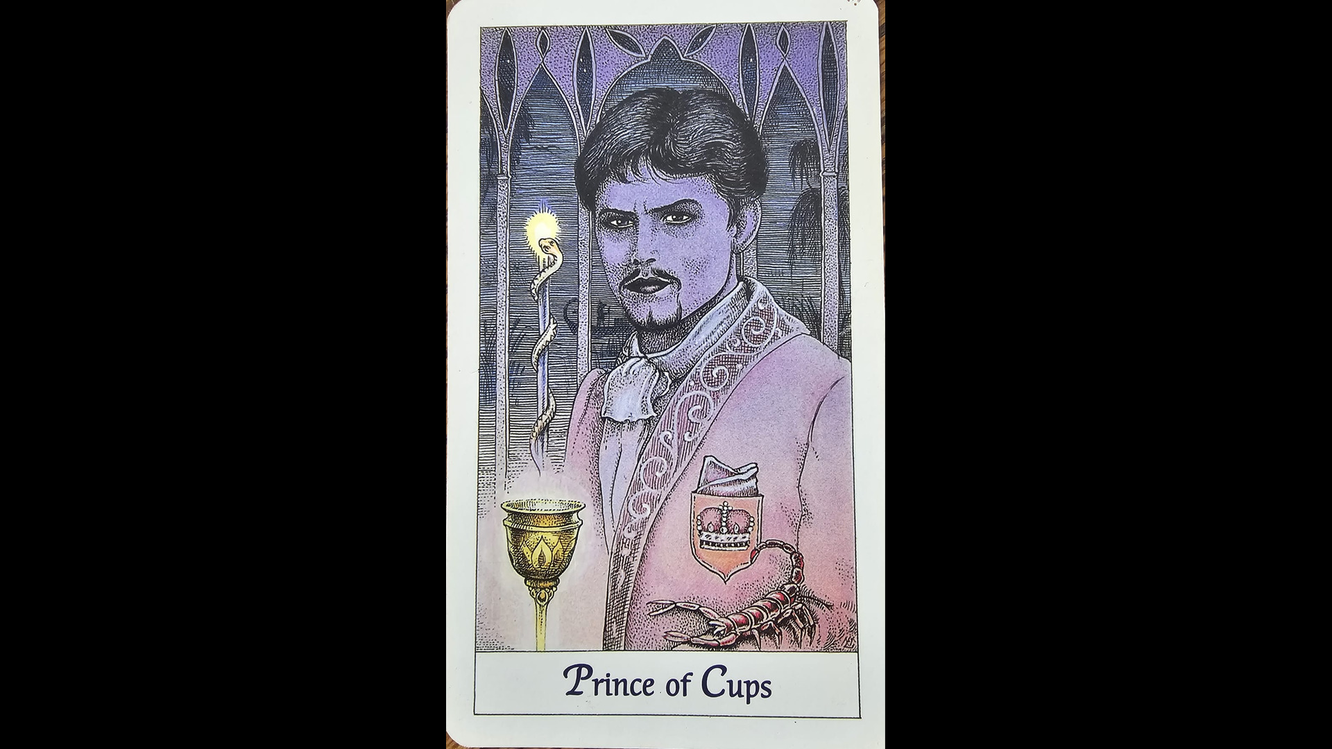 Prince of Cups tarot card on a black background: a prince raises a golden cup, a serpent rises from it and a scorpion rests on his arm.