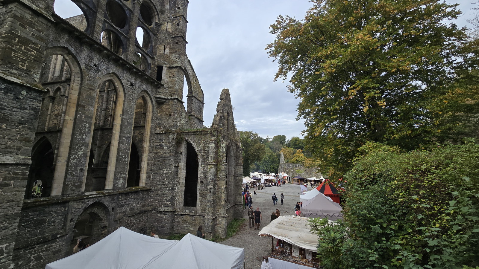 Gothic ruined abbey walls and empty arched windows frame a tree and a gravel courtyard where visitors walk among white market tents.